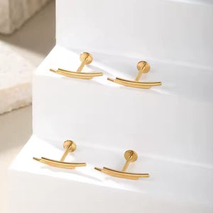 "Quatro piercings modelo labret na cor dourada, dispostos sobre degraus brancos iluminados. As joias são confeccionadas em Titânio G23 e possuem haste reta com base plana. O topo decorativo é minimalista, formado por duas barras lisas e paralelas levemente curvadas. Joia geométrica ideal para perfuração em conch, flat e helix."