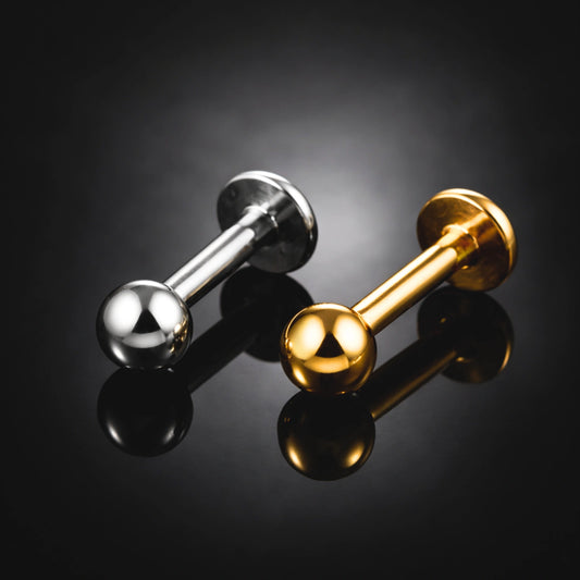 "Dois piercings modelo labret básico com topo de bolinha descansando sobre uma superfície preta reflexiva. Uma peça é na cor prateada natural do titânio e a outra na cor dourada. Ambos possuem haste reta lisa, base plana e sistema de rosca interna. Joia de Titânio G23 ideal para perfuração profissional no tragus, helix e boca."