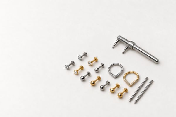 Joias de piercing em titânio, incluindo labrets e argolas clicker, ao lado de um Piercímetro profissional sobre fundo claro.