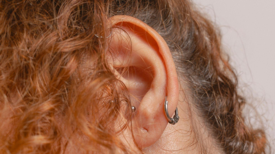 A orelha apresenta múltiplos piercings, incluindo um argola no lóbulo e o que parece ser um piercing no hélice.