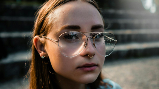 Retrato frontal de uma jovem usando óculos de grau grandes com gotas de água nas lentes. Ela possui um piercing de argola no septo nasal e brincos pequenos no lobo, com uma iluminação suave de fim de tarde ao fundo.