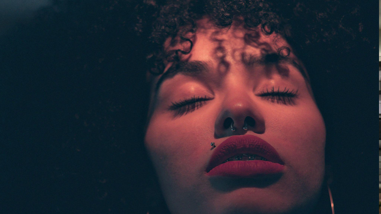 A foto é um close-up do rosto de uma mulher, capturado de um ângulo inferior (low angle). A iluminação é dramática e quente, com tons avermelhados e rosados que banham sua pele, criando sombras profundas que realçam os contornos do rosto.