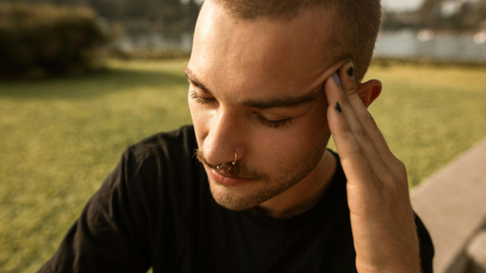 Esta imagem apresenta um retrato em close de um homem jovem em um ambiente externo ao ar livre. Ele parece estar desconfortável.