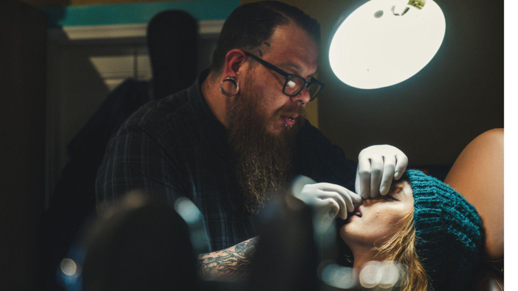 Um profissional usando luvas descartáveis trabalha com atenção no rosto de uma mulher deitada, que está reclinada em uma maca e coberta por um tecido.