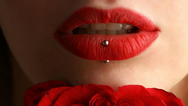 Lábios femininos pintados com um batom vermelho vibrante e levemente abertos, revelando os dentes. Há um piercing vertical no lábio inferior (labret).