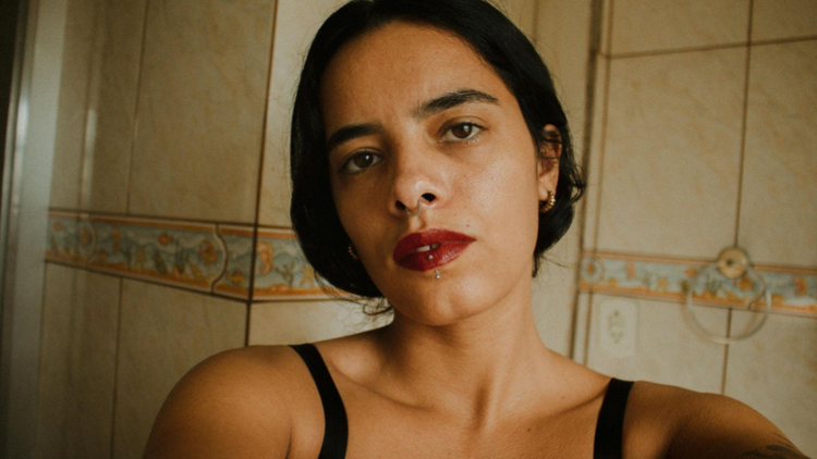Selfie de uma mulher de pele parda em um ambiente interno com azulejos bege ao fundo. Ela tem cabelos pretos presos e usa batom vinho escuro.