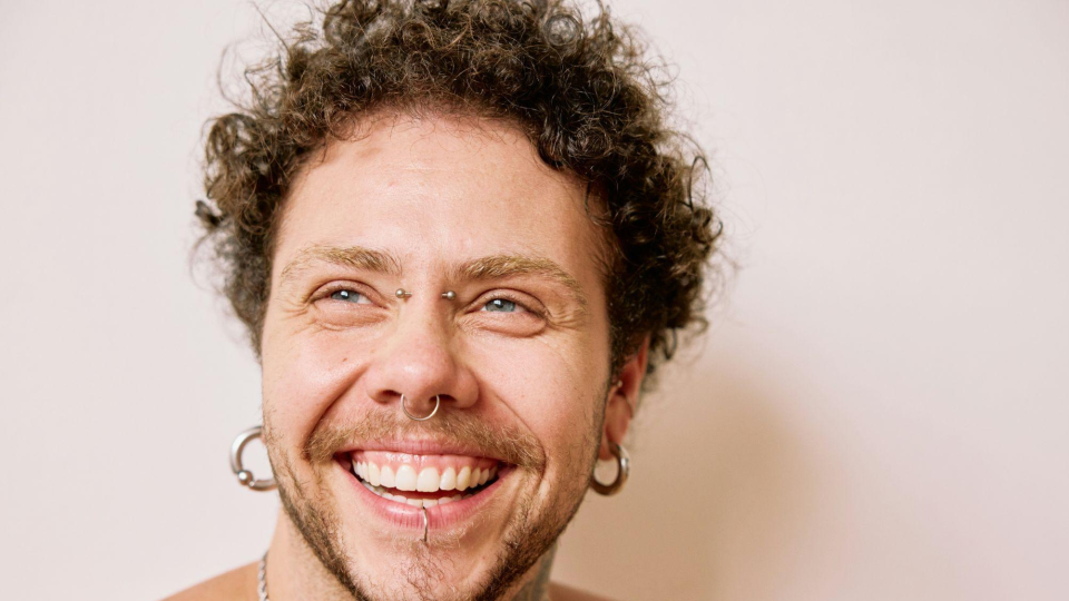 Imagem mostra um jovem sorrindo. Ele tem piercings prateados nos olhos, orelhas, nariz e lábios