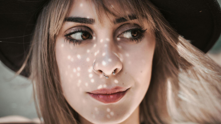 Uma imagem de estética suave onde uma mulher jovem, usando um chapéu de abas largas, recebe pontos de luz solar no rosto. Ela possui dois piercings no nariz: uma argola fina na narina e um piercing de septo do tipo ferradura.