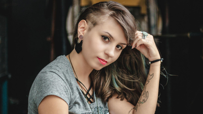 Uma mulher jovem com um corte de cabelo undercut (lateral raspada) e cabelos castanhos longos. Ela usa várias joias: um alargador na orelha, um brinco preto pendente, um piercing no lábio inferior e anéis de prata.
