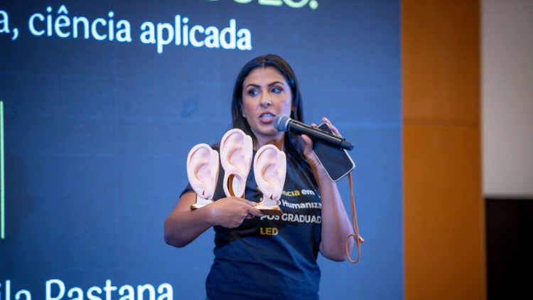 A imagem mostra uma palestrante em um evento ou aula especializada, apresentando conteúdos relacionados à área de piercing e anatomia aplicada.
