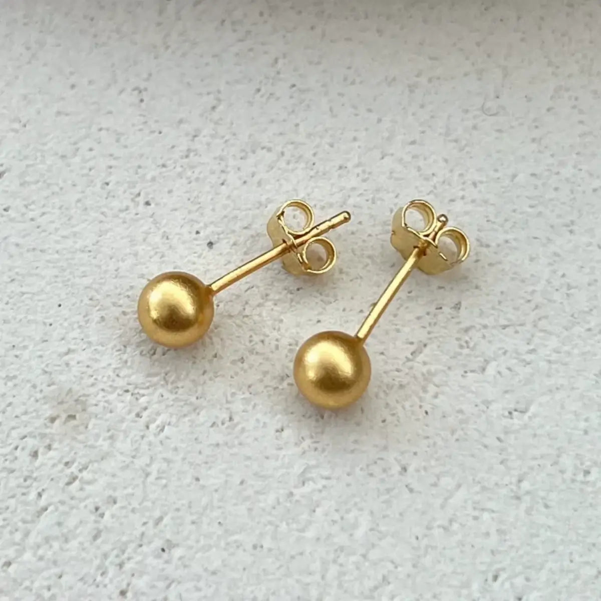 Par de brincos metálicos de aço cirúrgico cor dourada em formato de bolinha sobre superfície clara