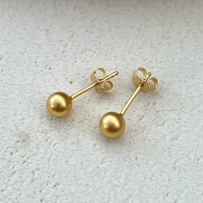 Par de brincos metálicos de aço cirúrgico cor dourada em formato de bolinha sobre superfície clara