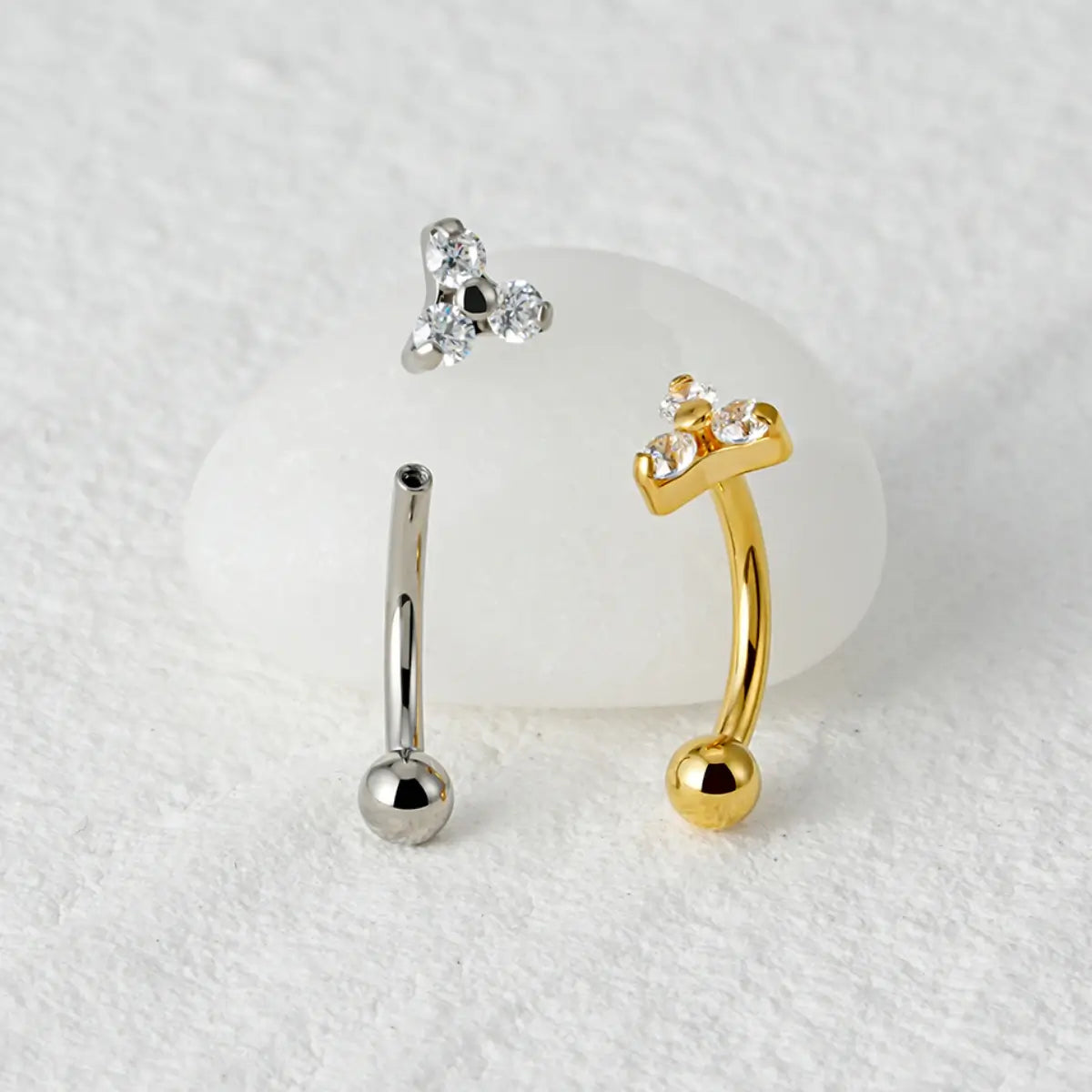 Piercings curvos banana de titânio prateado e dourado com 3 zircônias sobre superfície de mármore claro