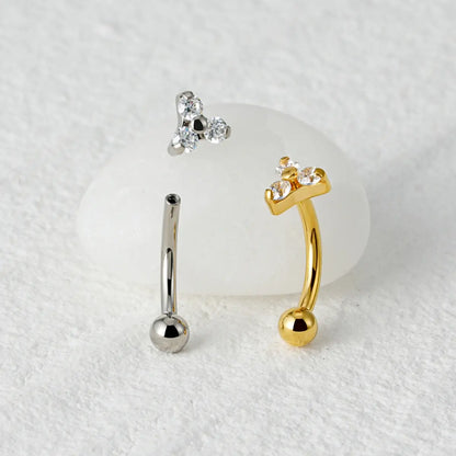 Piercings curvos banana de titânio prateado e dourado com 3 zircônias sobre superfície de mármore claro