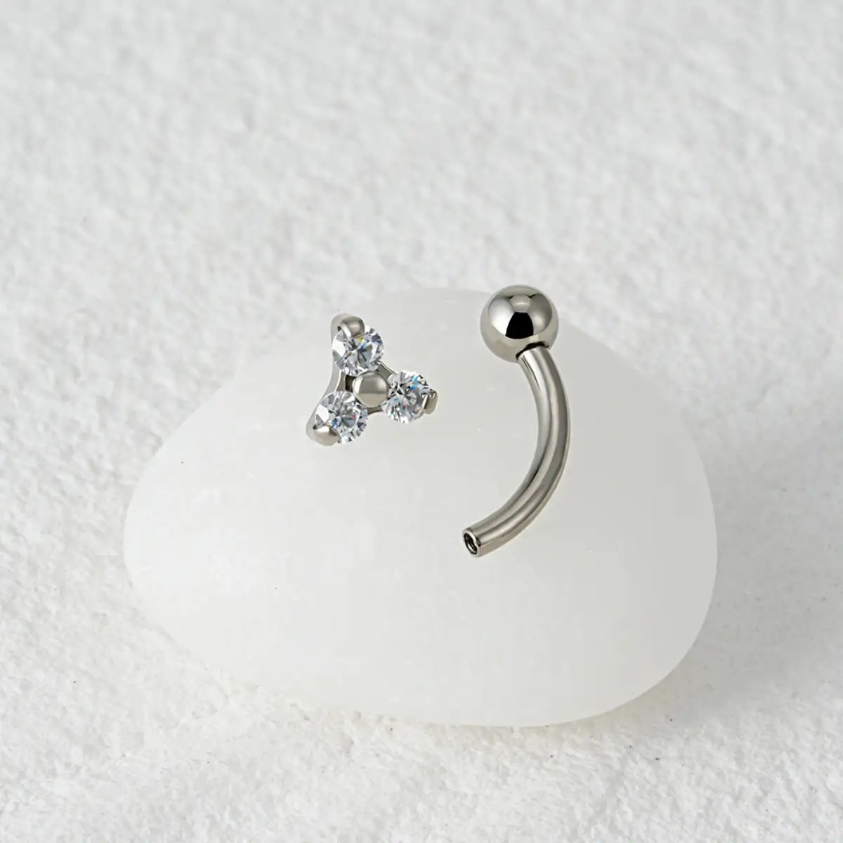 Piercing curvo banana prateado de titânio com 3 zircônias em detalhe sobre fundo de mármore