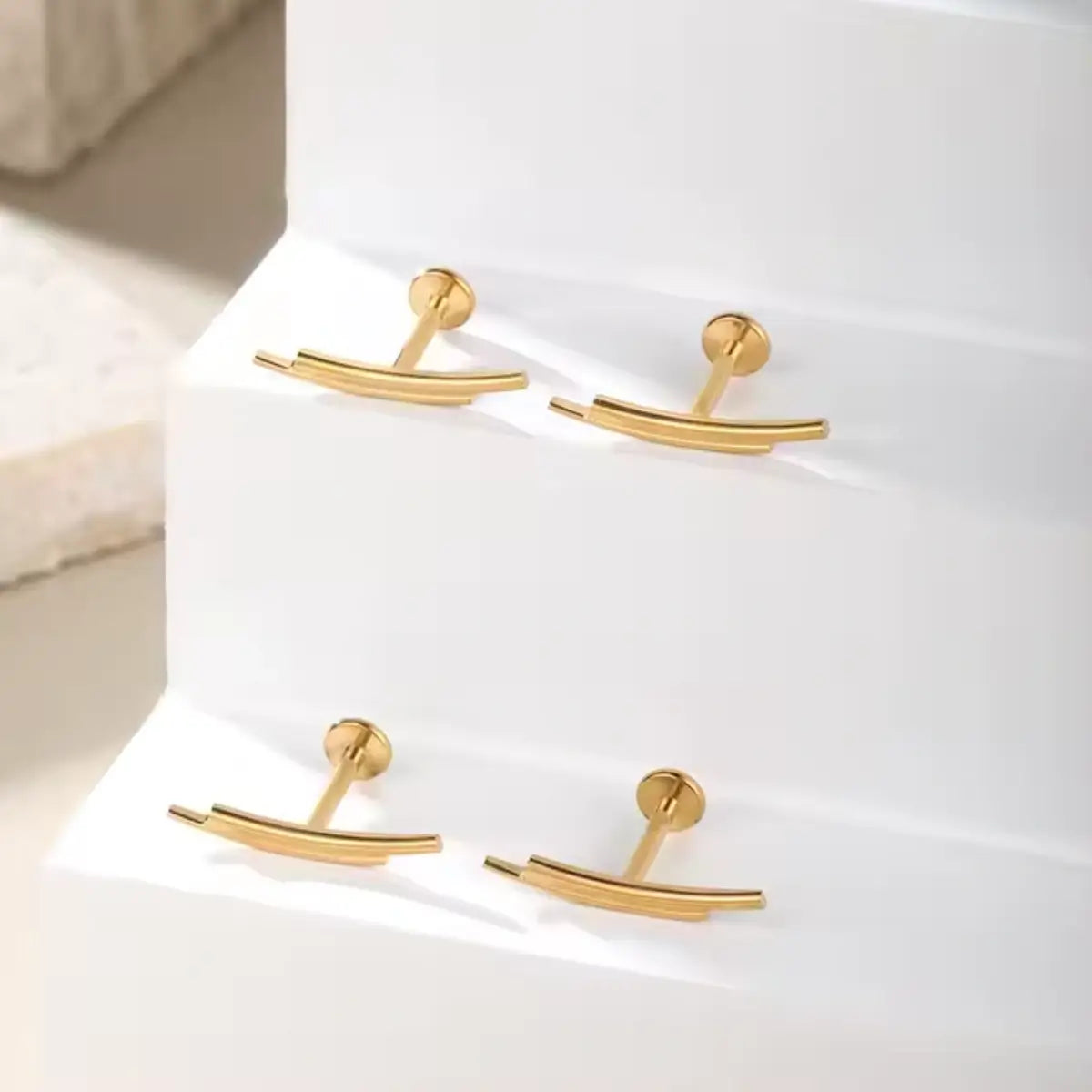 "Quatro piercings modelo labret na cor dourada, dispostos sobre degraus brancos iluminados. As joias são confeccionadas em Titânio G23 e possuem haste reta com base plana. O topo decorativo é minimalista, formado por duas barras lisas e paralelas levemente curvadas. Joia geométrica ideal para perfuração em conch, flat e helix."