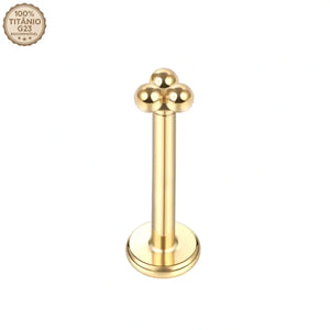 Joia de piercing modelo labret dourado feito em Titânio G23. Possui haste reta com base plana e topo decorativo em formato de cluster com 3 bolinhas lisas. No canto superior esquerdo há um selo de qualidade escrito 100% Titânio G23 Biocompatível. Ideal para tragus, helix e perfuração humanizada.