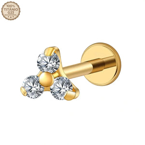 "Joia de piercing modelo labret dourado confeccionada em Titânio G23. Apresenta haste reta com base plana e topo decorativo com três pedras de zircônia redondas e brilhantes agrupadas em formato de triângulo. No canto superior esquerdo da imagem, há um selo atestando '100% Titânio G23 Biocompatível'. Ideal para perfurações em tragus, conch e helix."