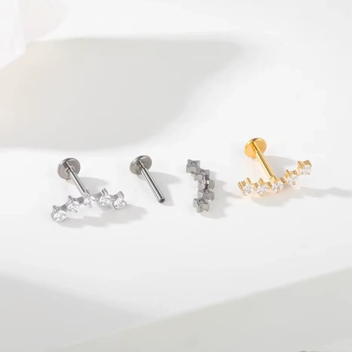 Variações de piercings labret de titânio com cluster espaçado de 5 zircônias nas cores prata e dourado sobre fundo claro