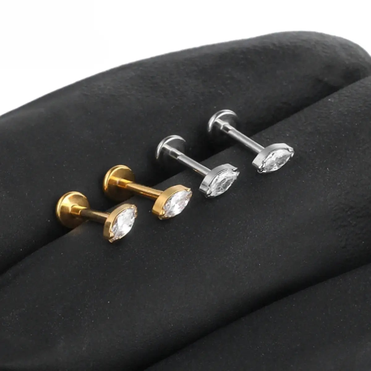 "Quatro piercings modelo labret dispostos lado a lado sobre uma superfície escura texturizada. Dois piercings são na cor dourada e dois na cor prateada. Todos são feitos em Titânio G23 e possuem haste reta com base plana e uma pedra de zircônia brilhante com lapidação navete (marquise) no topo. Joia ideal para perfuração humanizada no tragus, medusa e helix."
