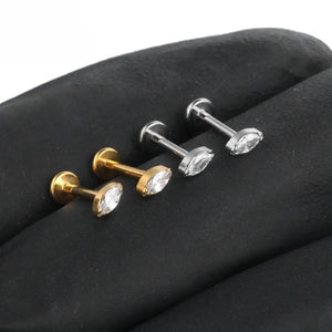 "Quatro piercings modelo labret dispostos lado a lado sobre uma superfície escura texturizada. Dois piercings são na cor dourada e dois na cor prateada. Todos são feitos em Titânio G23 e possuem haste reta com base plana e uma pedra de zircônia brilhante com lapidação navete (marquise) no topo. Joia ideal para perfuração humanizada no tragus, medusa e helix."