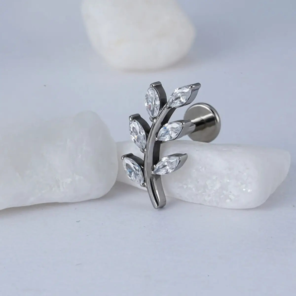 Piercing labret de titânio prata design ramo com 6 zircônias em formato de folhas sobre fundo claro com pedras decorativas