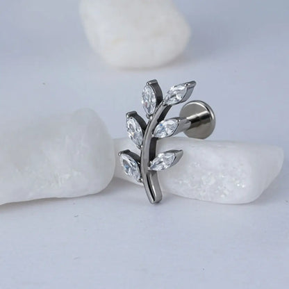 Piercing labret de titânio prata design ramo com 6 zircônias em formato de folhas sobre fundo claro com pedras decorativas